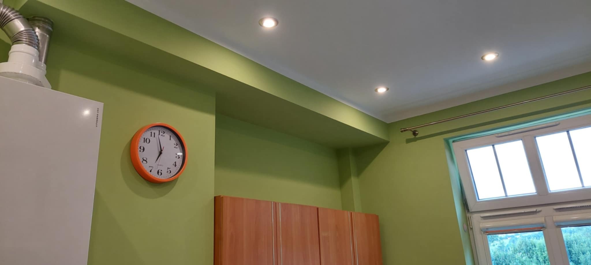 Ściana w odcieniach zieleni z pomarańczowym zegarem, oświetleniem punktowym i fragmentem białej szafki. Widoczne okno z roletami i fragment mebli.
