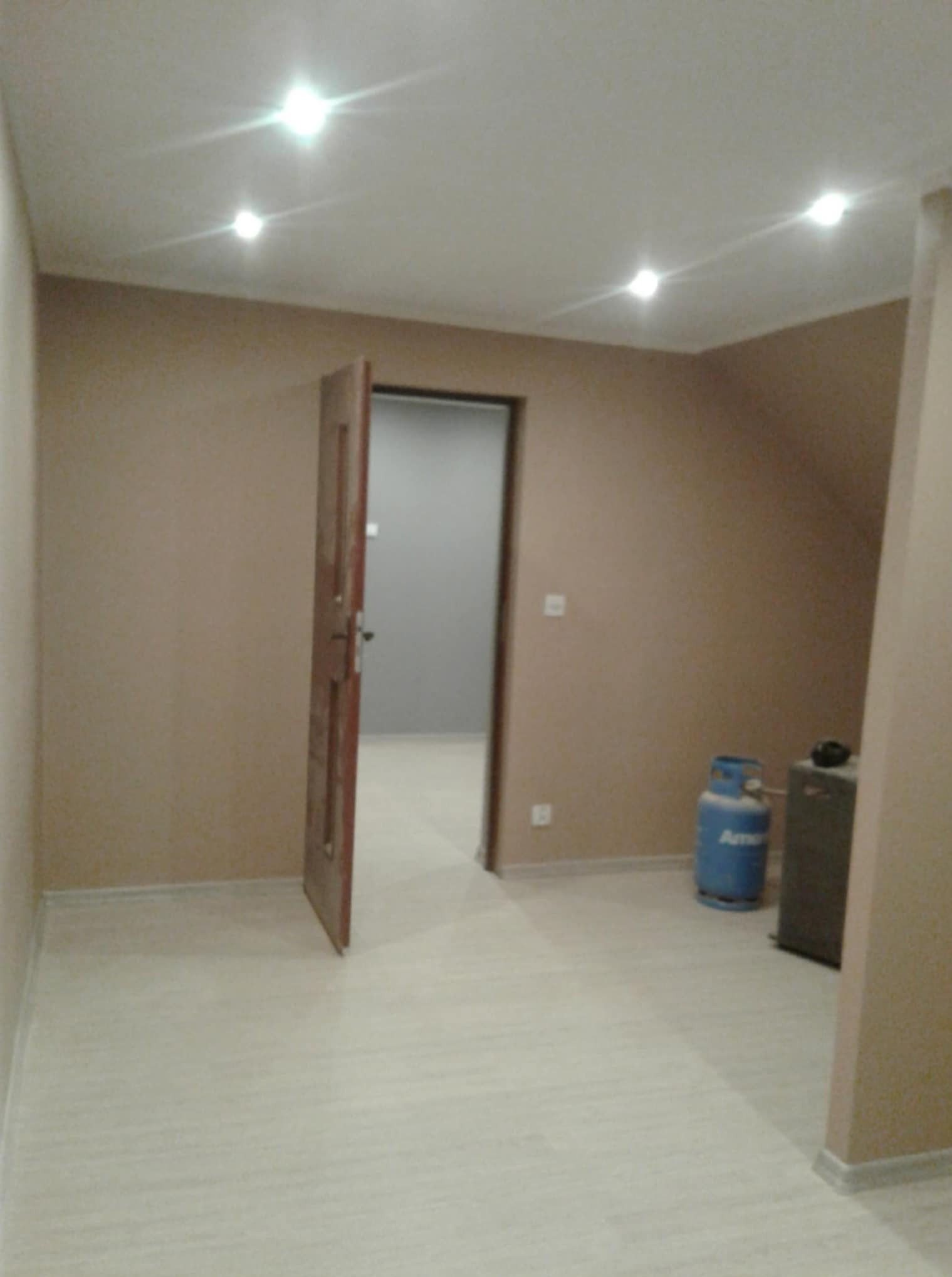 Wnętrze pokoju po remoncie: beżowe ściany, jasna podłoga, otwarte drewniane drzwi. Widoczna butla gazowa i sprzęt. Oświetlenie sufitowe punktowe.