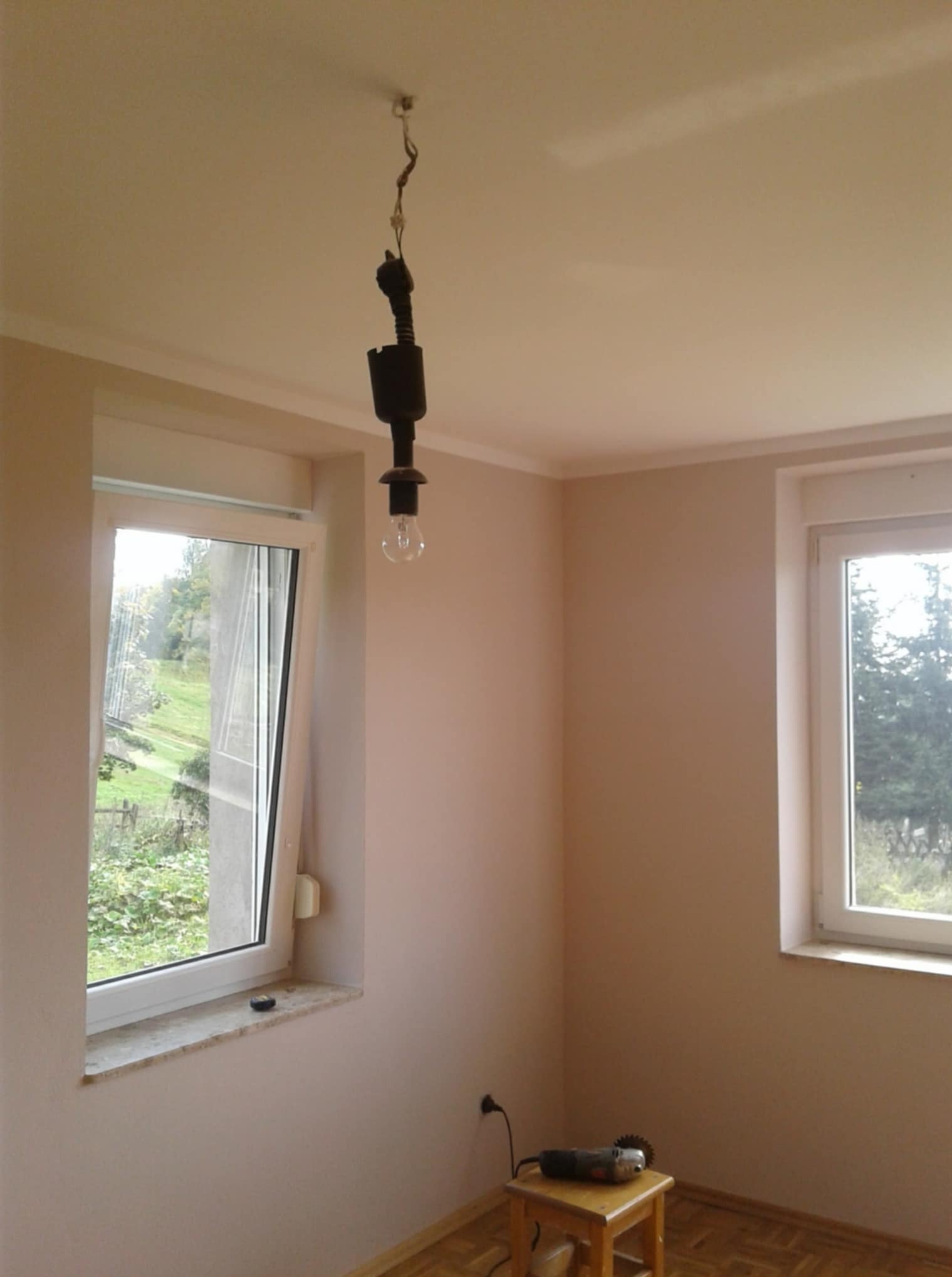 Pokój po remoncie w Nowej Rudzie: świeżo pomalowane ściany w odcieniach beżu, dwa okna, wisząca żarówka i narzędzia na taborecie, gotowe do dalszych prac.