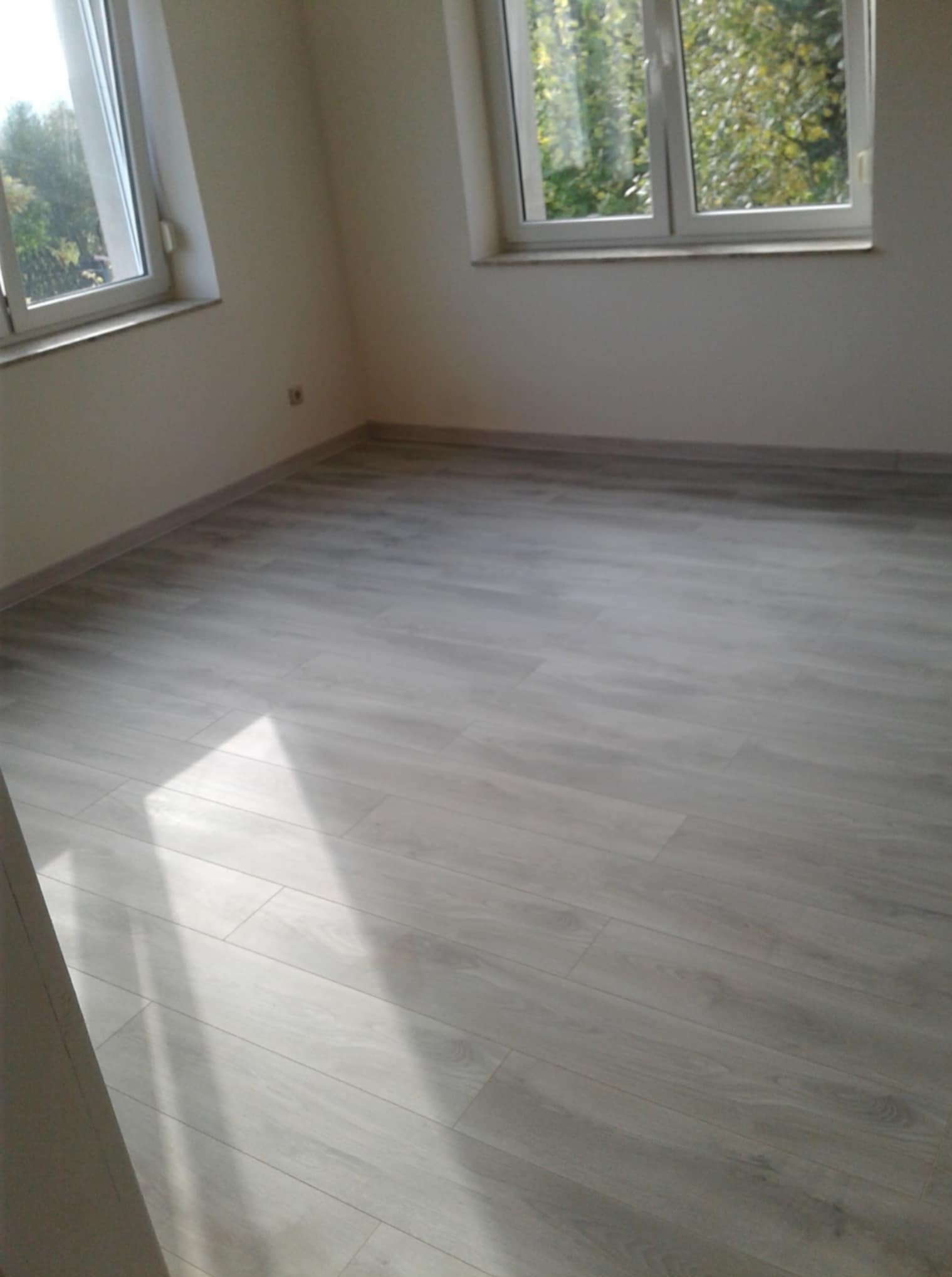 Jasne panele podłogowe w pustym pokoju z dwoma oknami, widok na drzewa. Świeżo wykończone wnętrze po remoncie, gotowe do umeblowania.