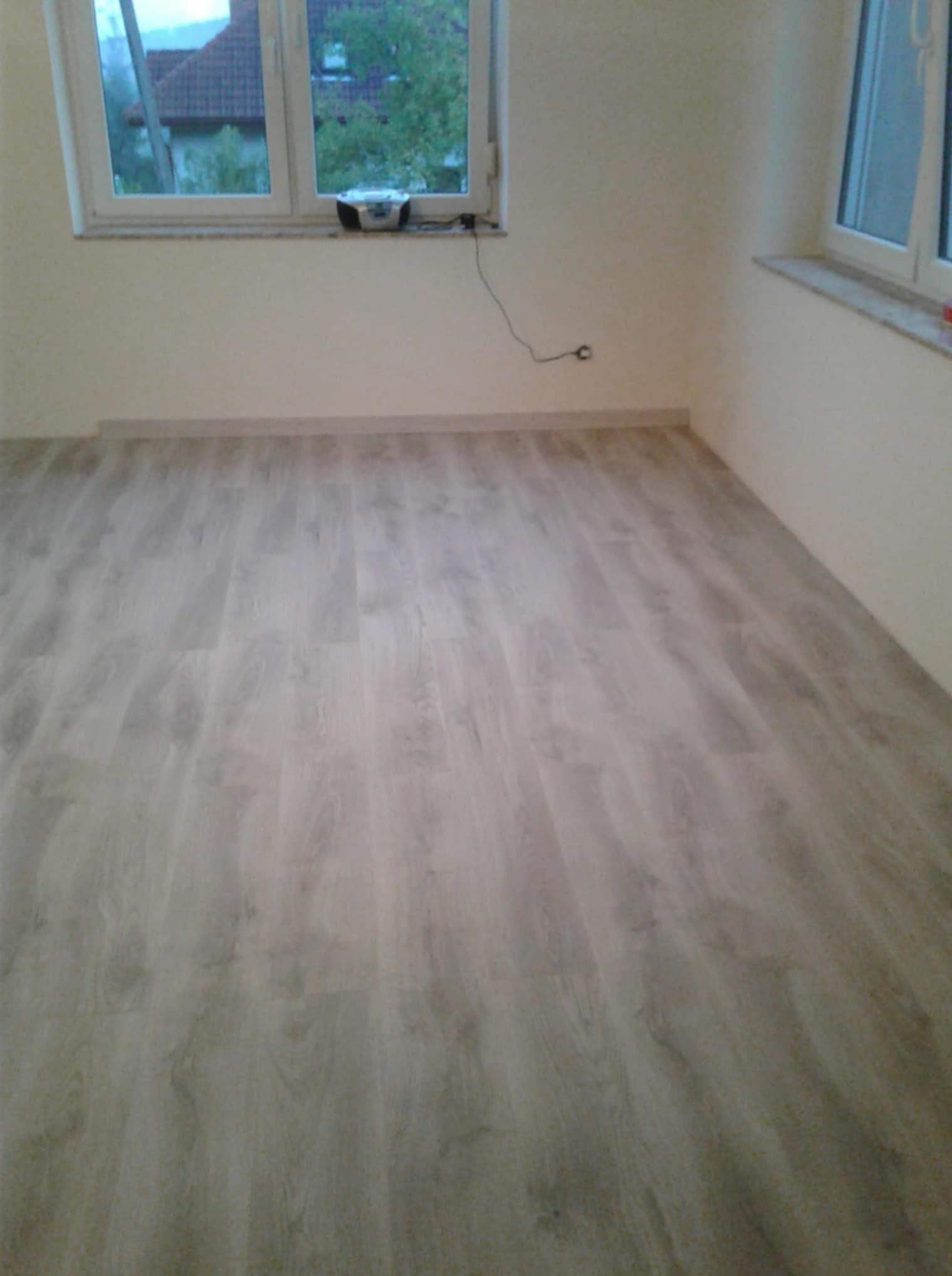 Jasne panele podłogowe w pokoju z oknami, widok na zewnątrz. Na parapecie stoi radio. Ściany w kolorze ciepłej bieli.