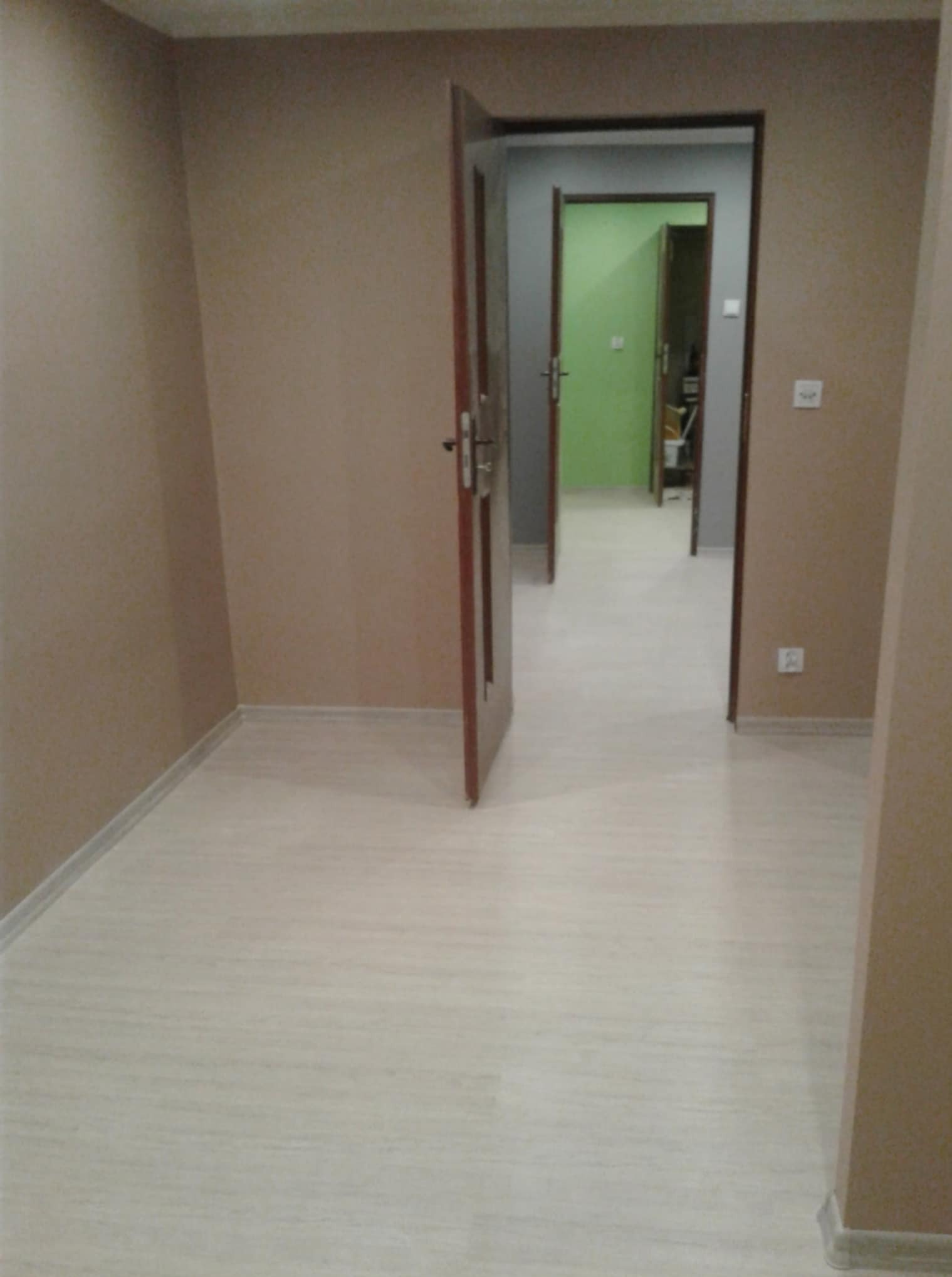 Jasne panele podłogowe w pustym pokoju z beżowymi ścianami. Otwarte drzwi ukazują dalszą część mieszkania z zielonymi ścianami.