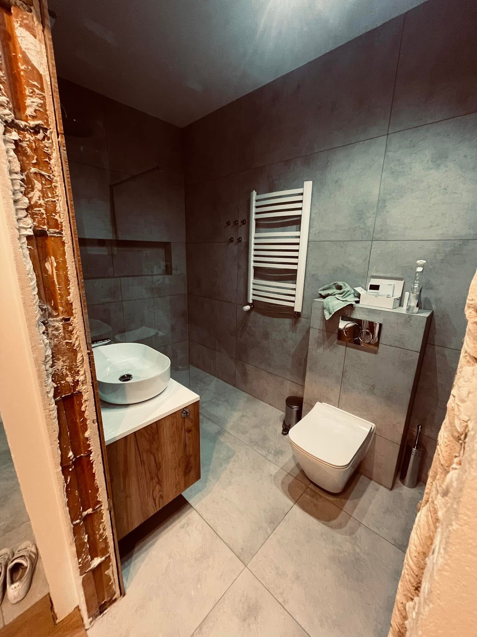 Nowoczesna łazienka w szarościach z drewnianymi akcentami, umywalka nablatowa i grzejnik drabinkowy. Widoczne ślady po demontażu ościeżnicy drzwiowej.