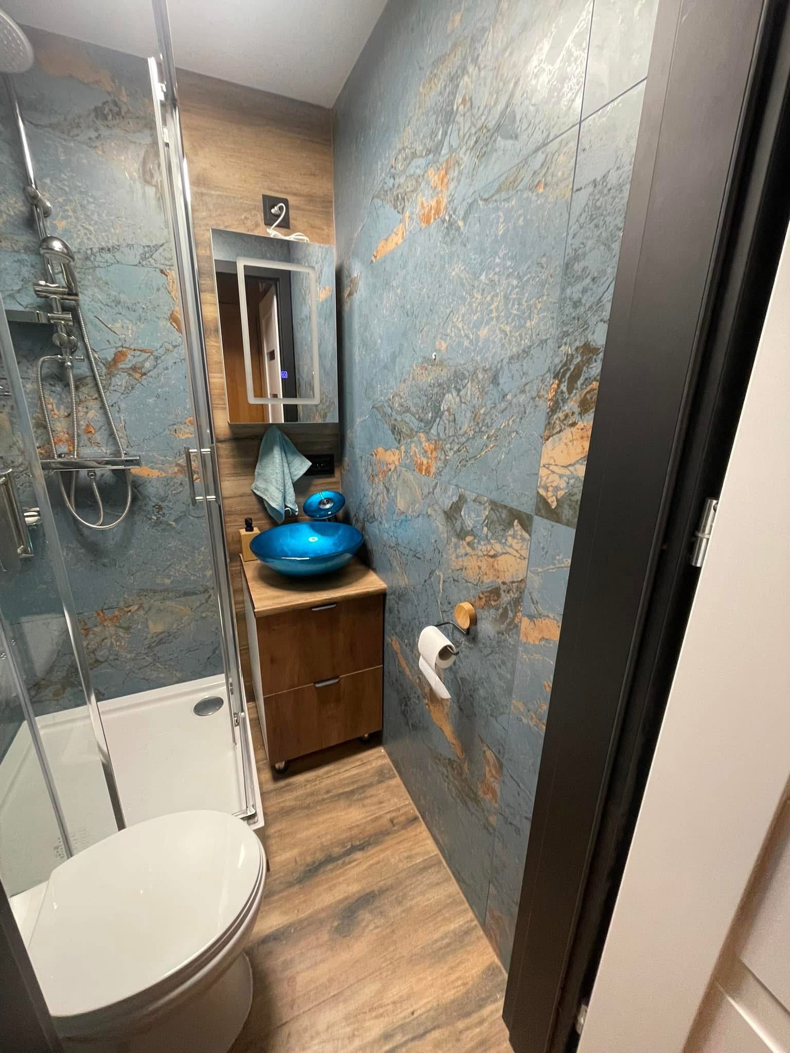 Wykończona łazienka z kabiną prysznicową, toaletą, szafką z umywalką w kolorze niebieskim i płytkami imitującymi marmur na ścianach.