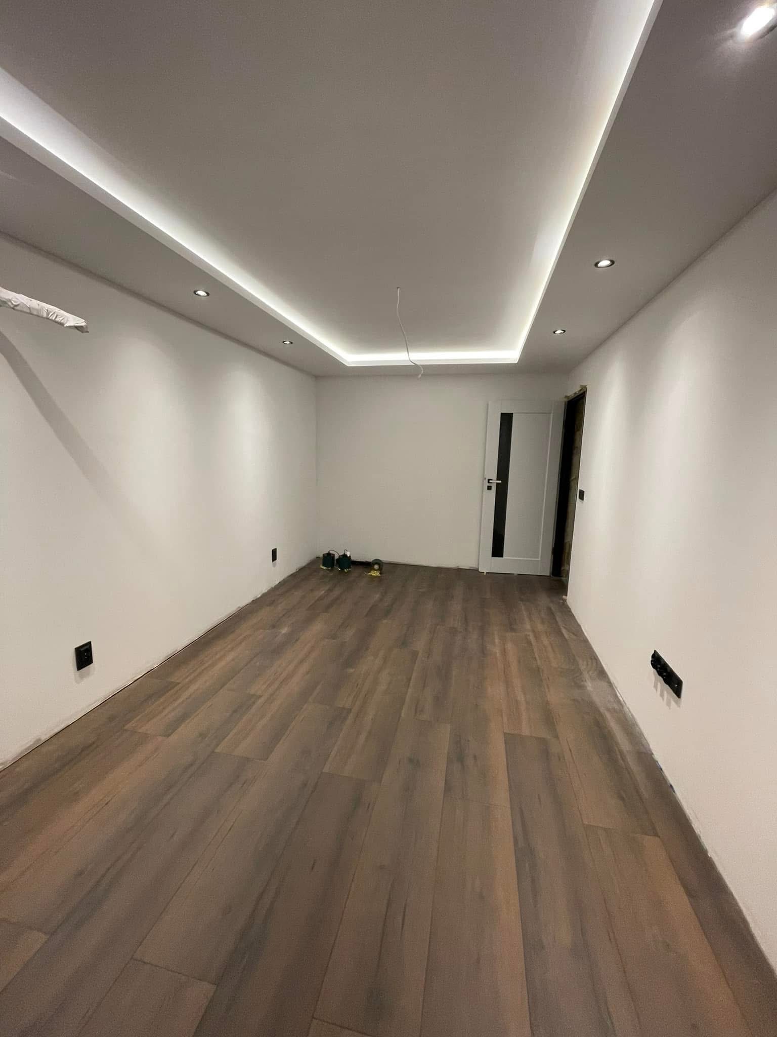 Jasne wnętrze po remoncie: panele podłogowe, białe ściany, sufit z oświetleniem LED i zamontowane drzwi z czarnym akcentem. Widoczne gniazdka i narzędzia.