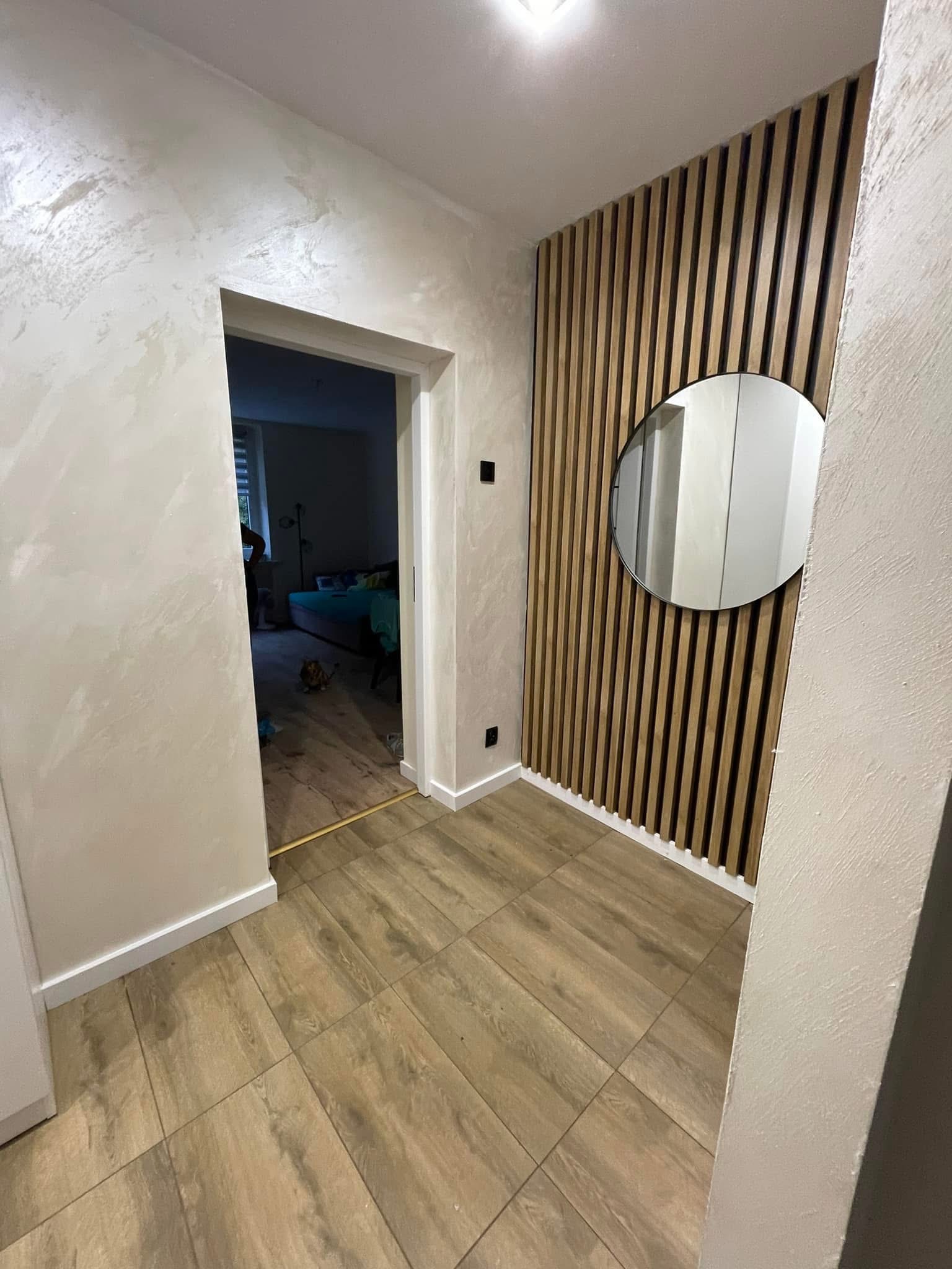 Korytarz po remoncie: dekoracyjna ściana z drewnianych lameli z okrągłym lustrem, ozdobne wykończenie ścian, podłoga z płytek imitujących drewno i widok na pokój.