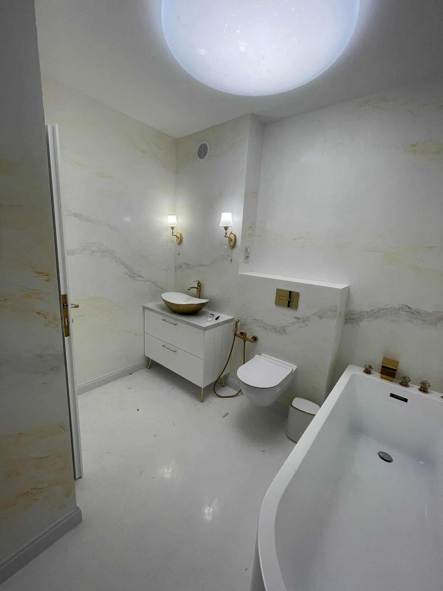 Elegancka łazienka w bieli i złocie: umywalka na szafce, toaleta podwieszana, wanna. Ściany zdobione wzorem marmuru, oświetlenie punktowe.