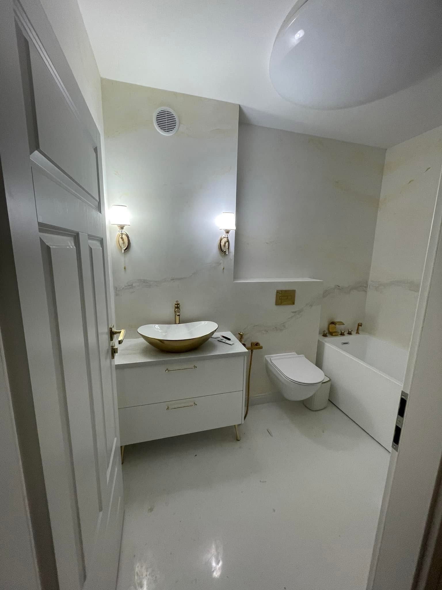 Nowoczesna łazienka w bieli i złocie: umywalka na szafce, toaleta podtynkowa, wanna z parawanem. Marmurowe wykończenie ścian, oświetlenie punktowe.