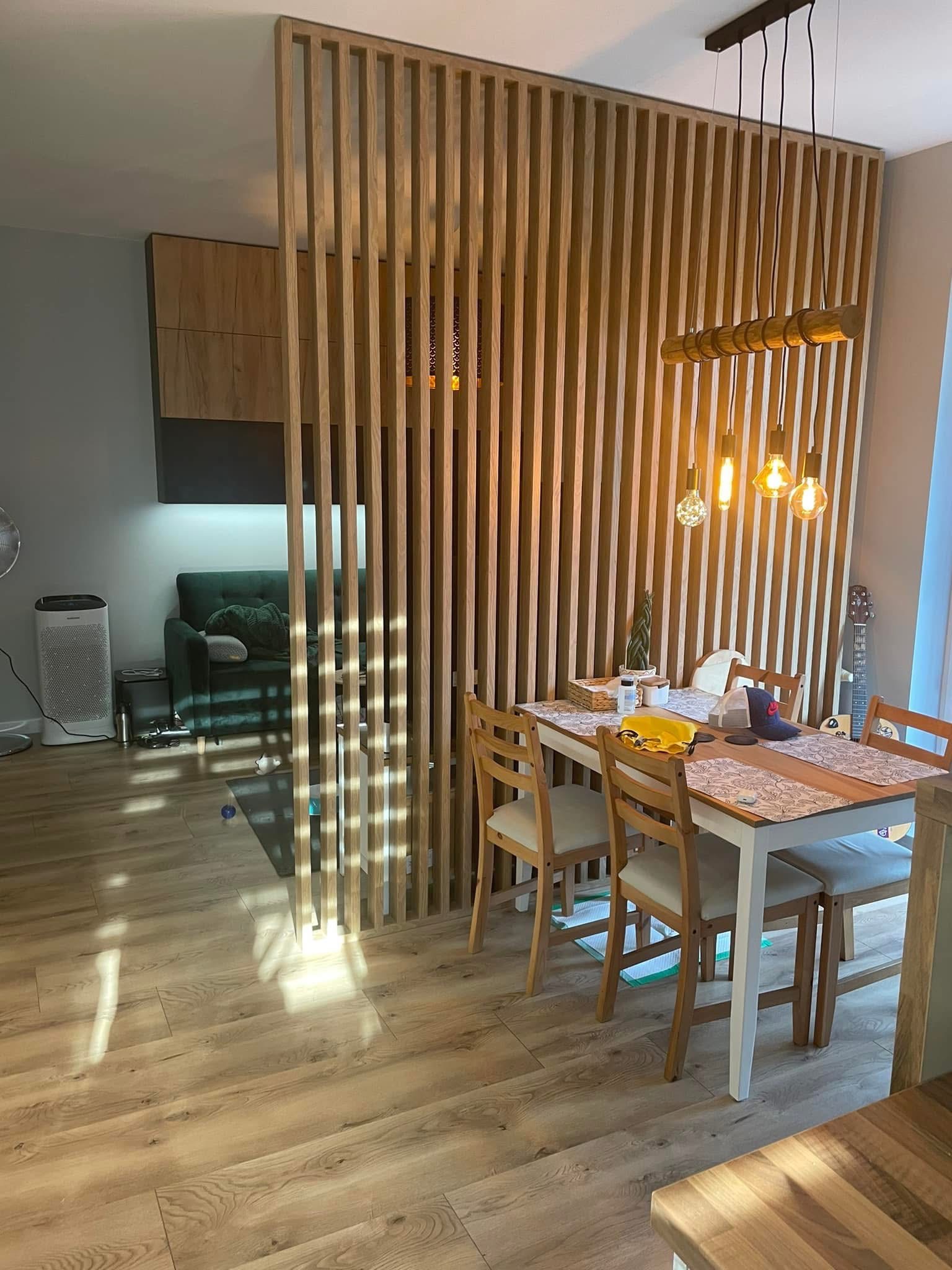 Drewniana ażurowa ścianka działowa oddziela salon od jadalni z drewnianym stołem i krzesłami. Nad stołem wisi lampa z żarówkami Edisona. Widoczna podłoga z paneli.