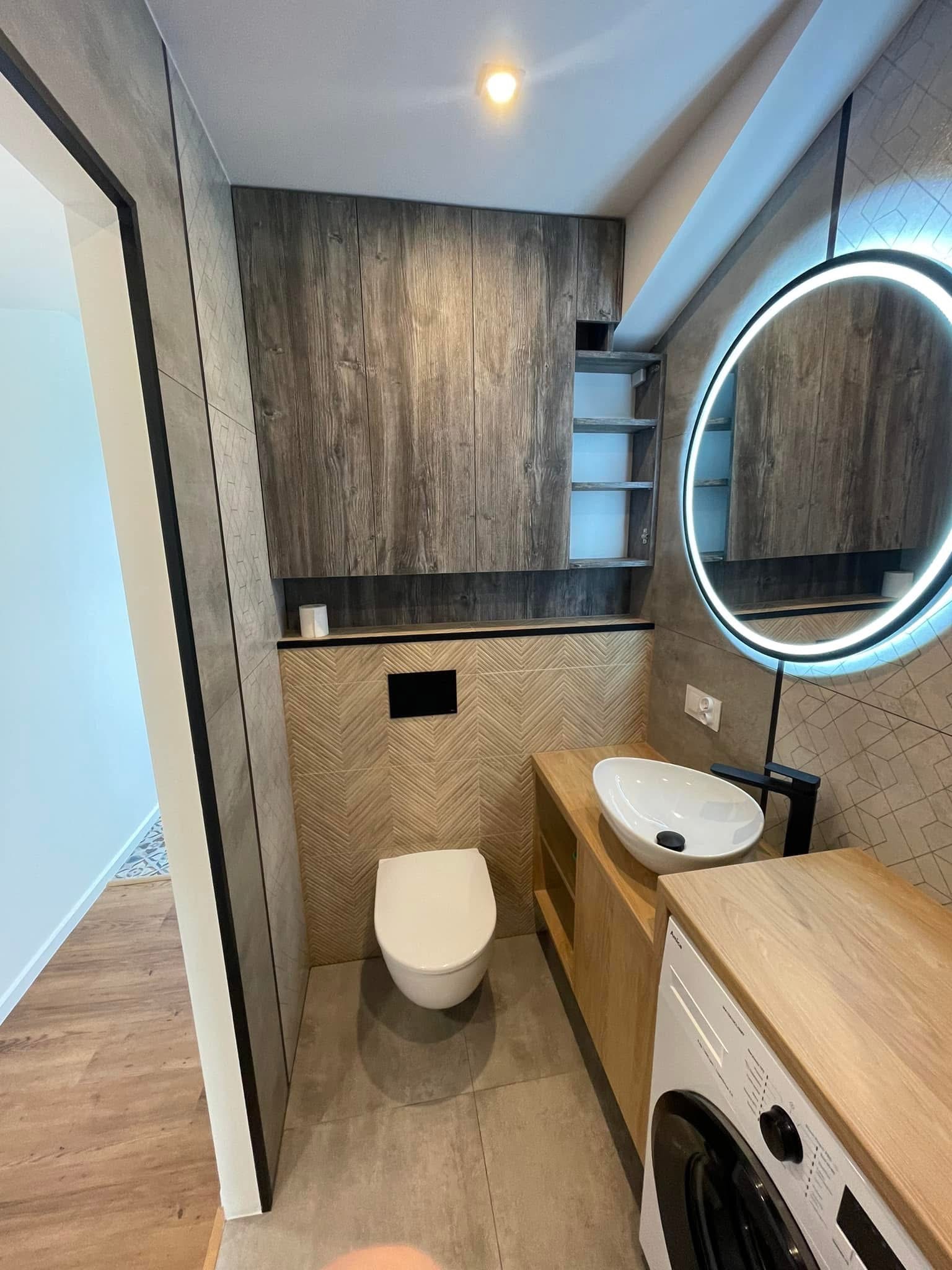 Nowoczesna łazienka z drewnianymi akcentami: szafki, blat pod umywalką i obudowa pralki. Umywalka nablatowa, okrągłe lustro z podświetleniem LED.
