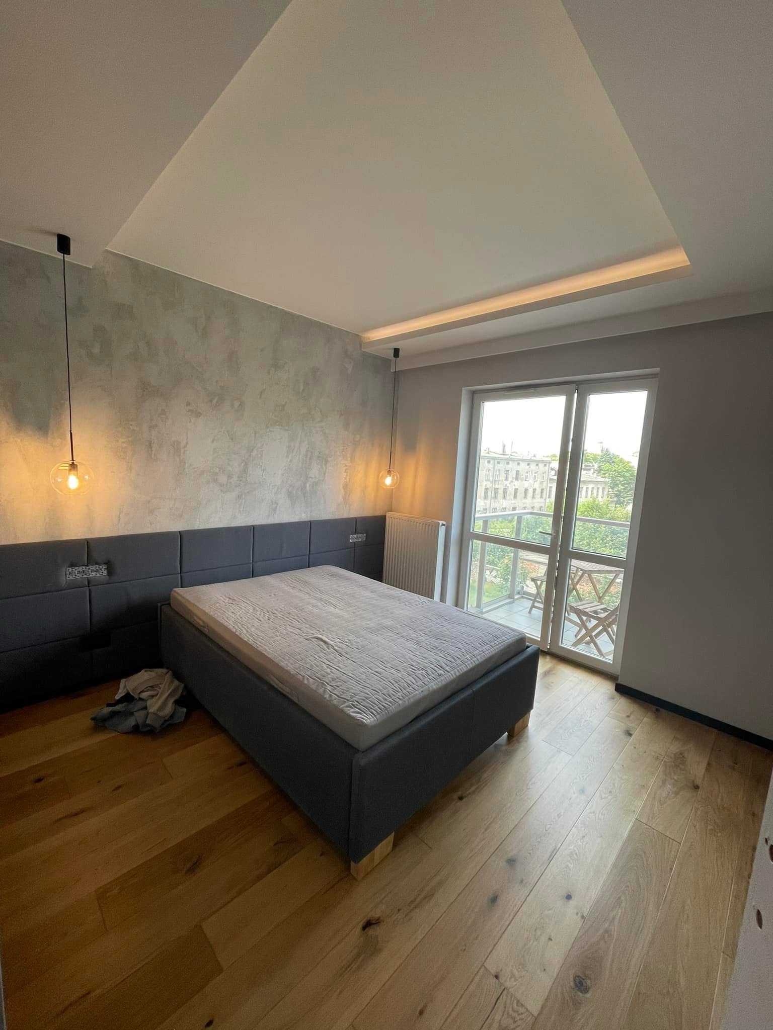 Nowoczesna sypialnia z łóżkiem, drewnianą podłogą, oświetleniem punktowym i wyjściem na balkon. Ściana pokryta dekoracyjnym tynkiem.