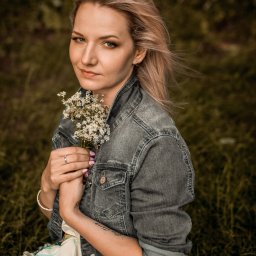 Czuły Kadr Fotografia - Portret kobiety z blond włosami, w dżinsowej kurtce, trzymającej bukiet polnych kwiatów na tle zieleni, z delikatnym uśmiechem i pierścionkiem na palcu.