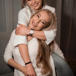 Czuły Kadr Fotografia - Uśmiechnięta blondynka obejmuje siedzącą kobietę w fotelu, obie ubrane w białe bluzki. Widoczny zegarek na ręce obejmującej osoby i szara zasłona w tle.