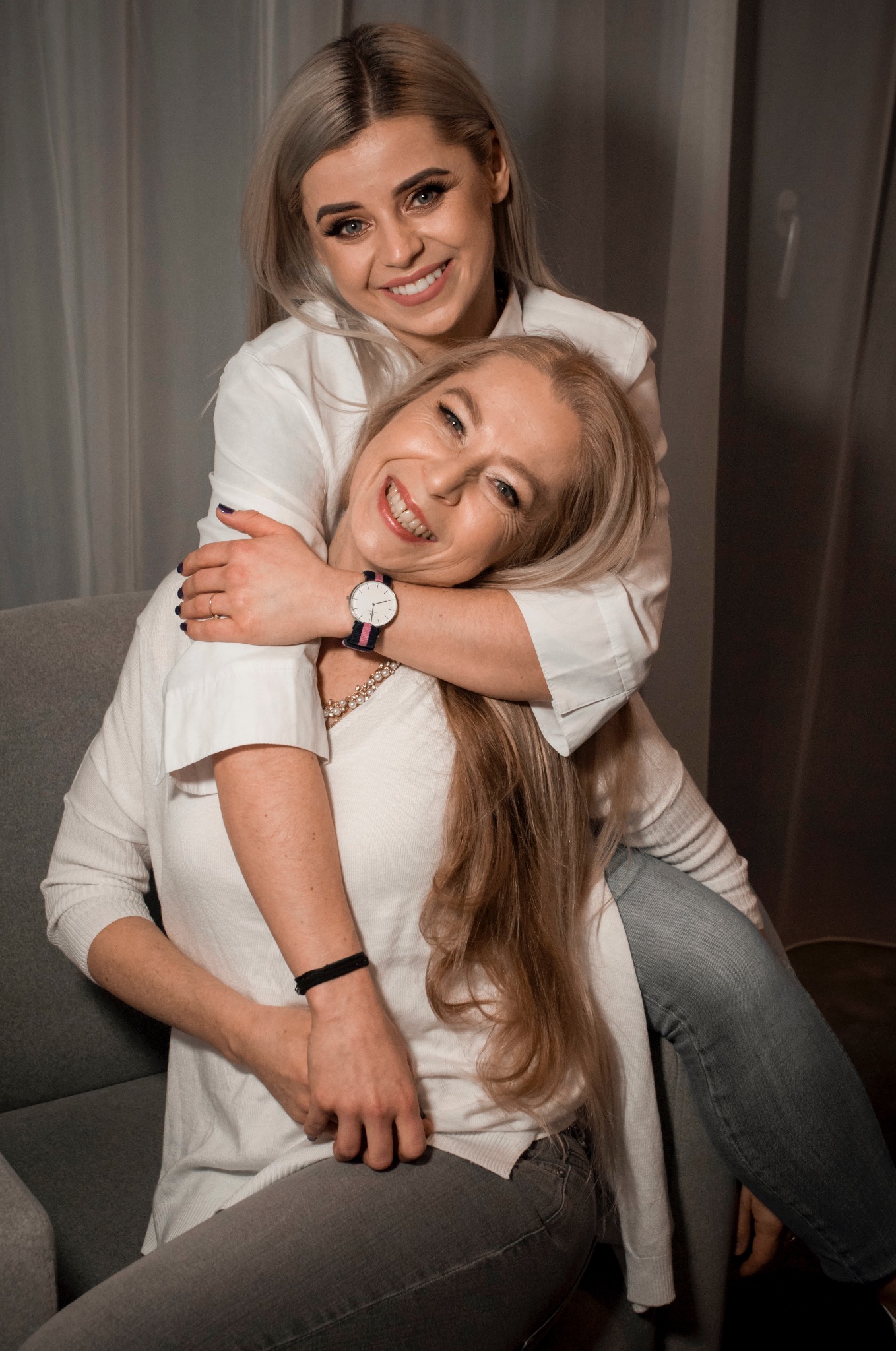 Uśmiechnięta blondynka obejmuje siedzącą kobietę w fotelu, obie ubrane w białe bluzki. Widoczny zegarek na ręce obejmującej osoby i szara zasłona w tle.
