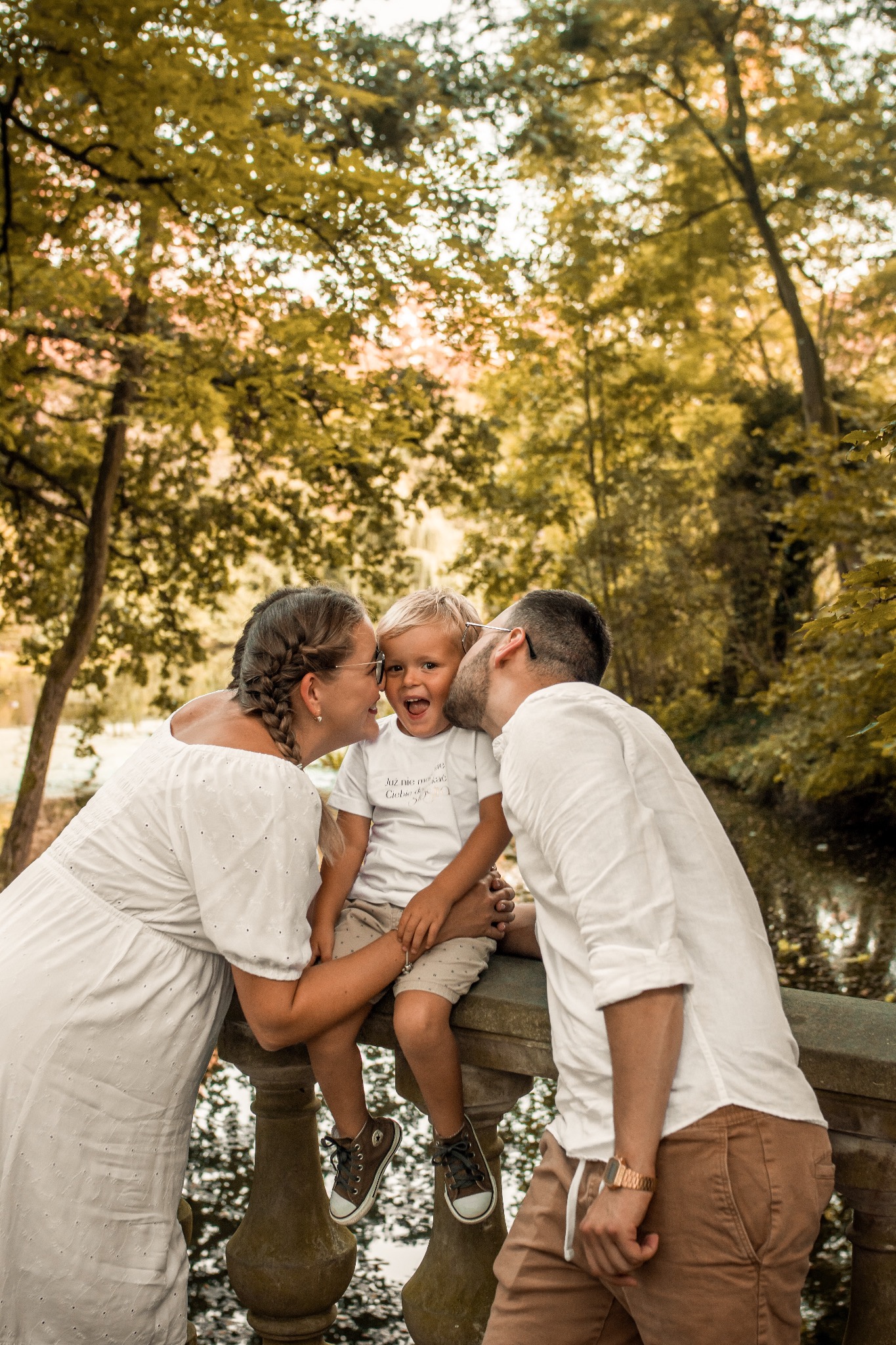 Uśmiechnięty chłopiec w objęciach rodziców, siedzący na kamiennej balustradzie mostu w parku. Mama i tata całują syna w policzki. Ciepłe, naturalne światło.