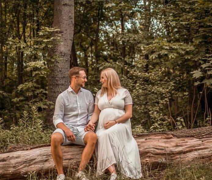 Ciążowa sesja w plenerze: przyszła mama w białej sukni i partner siedzą na pniu drzewa, trzymając się za ręce i patrząc na siebie z czułością, w tle leśny krajobraz.