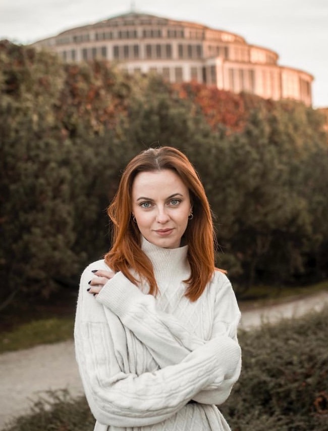 Portret kobiety w białym swetrze na tle Hali Stulecia w Chojnicach, wykonany podczas sesji plenerowej. Jesienne barwy drzew i miękkie światło.