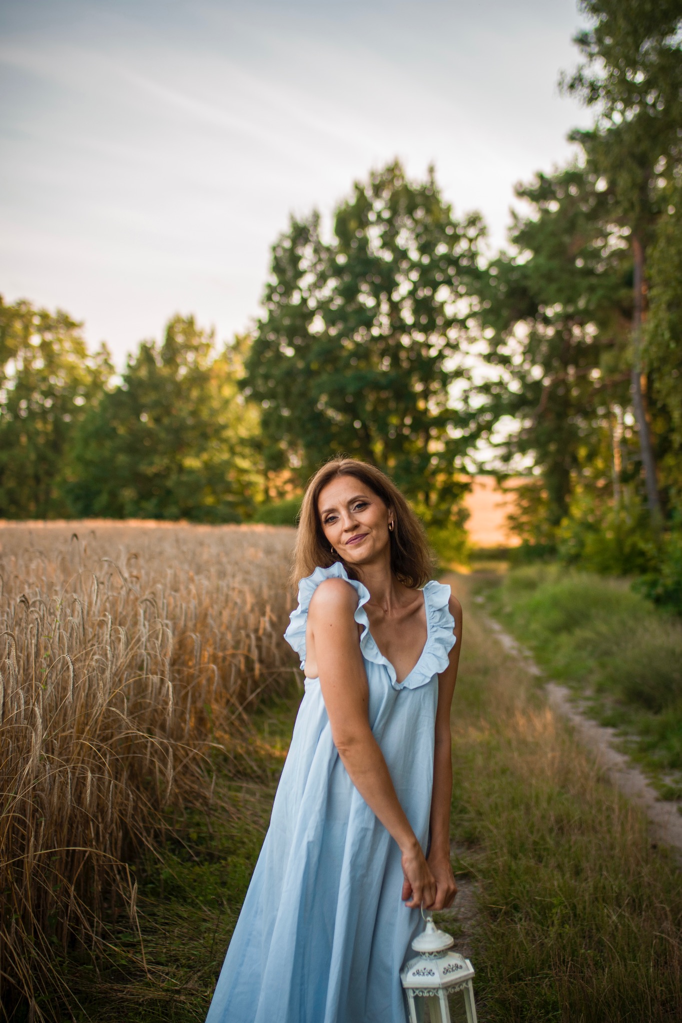 Portret kobiety w błękitnej sukience na tle pola zboża i drzew, trzymającej białą latarnię. Ujęcie w naturalnym świetle, z delikatnym uśmiechem.