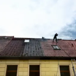 Mężczyzna czyści dach z czerwonej dachówki, widoczny efekt przed i po czyszczeniu. Na dole budynku żółta elewacja i drabina. Pochmurne niebo w tle.
