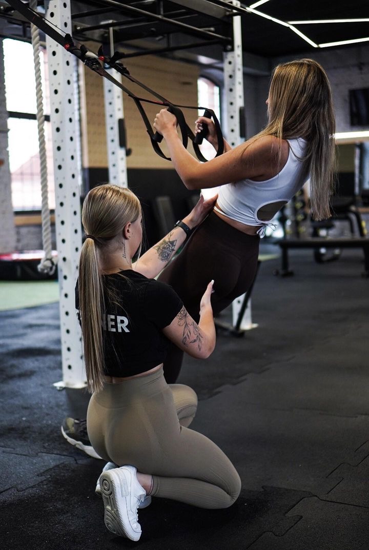 Trenerka personalna asekuruje klientkę podczas ćwiczeń na TRX w siłowni. Kobiety w sportowych strojach, widoczne tatuaże na ramionach trenerki.