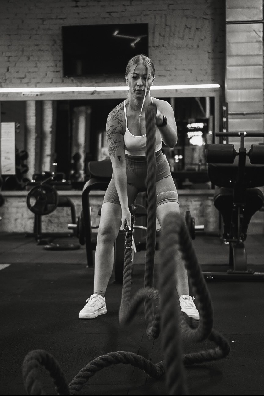 Kobieta z tatuażem ćwiczy z linami w siłowni, skupiona twarz, monochromatyczne ujęcie, widoczne wyposażenie siłowni w tle, dynamiczne ujęcie lin.
