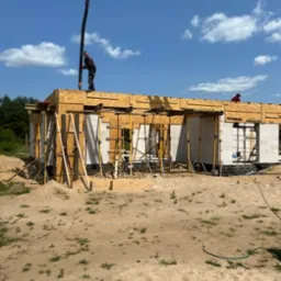 Budowa domu w trakcie: pracownik obsługuje pompę do betonu na szalunku stropu, ściany z bloczków, teren budowy z piaskiem. Słoneczny dzień.