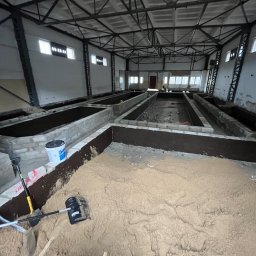 SCAVI SPÓŁKA Z OGRANICZONĄ ODPOWIEDZIALNOŚCIĄ - Wnętrze hali z przygotowanymi fundamentami pod budowę, widoczne narzędzia i materiały budowlane. Surowy stan, ceglane obrzeża, piasek.