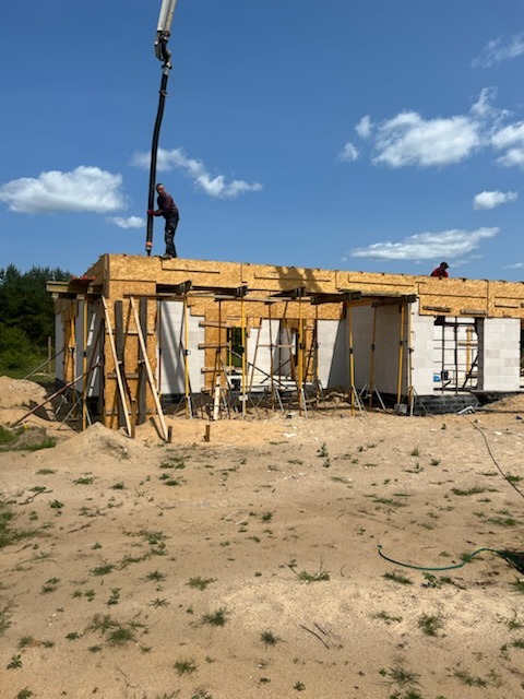 Budowa domu w trakcie: pracownik obsługuje pompę do betonu na szalunku stropu, ściany z bloczków, teren budowy z piaskiem. Słoneczny dzień.