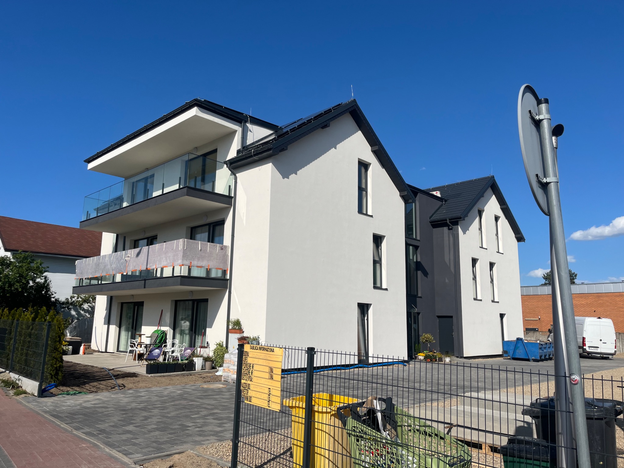 Nowoczesny, trzypiętrowy budynek z balkonami o szklanych balustradach, w trakcie wykończenia terenu. Widoczne śmietniki i tablica informacyjna budowy.