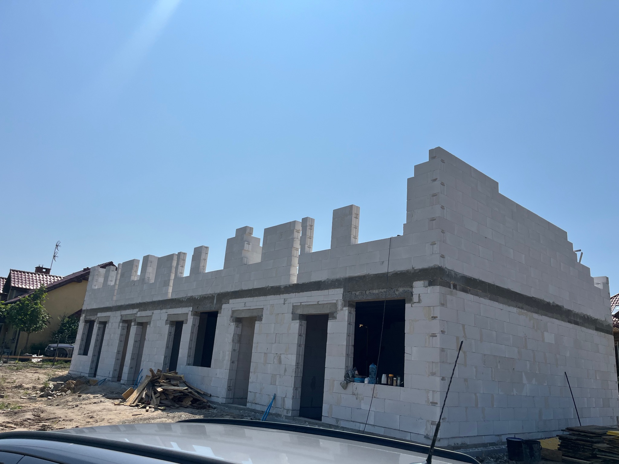 Wznoszenie ścian parterowego domu z białych bloczków, widoczne otwory okienne i drzwiowe, na pierwszym planie fragment dachu samochodu, słoneczny dzień.