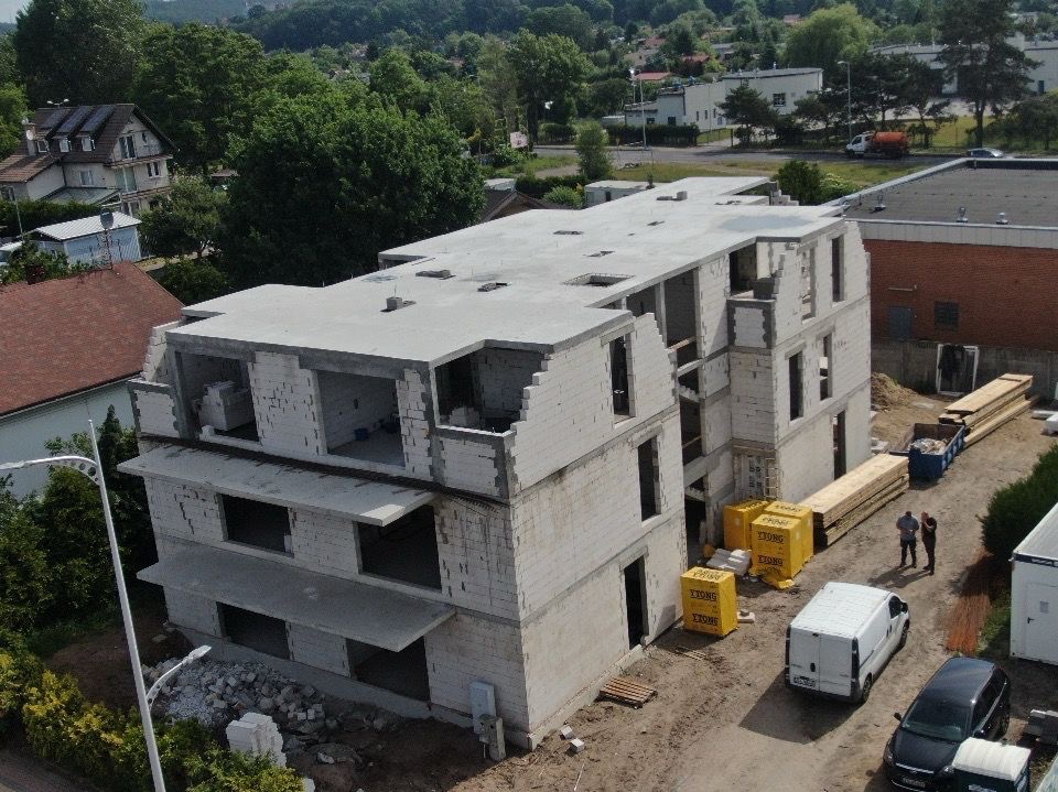 Wznoszenie trzypiętrowego budynku z betonu komórkowego Ytong, widok z góry. Częściowo otynkowany, z widocznymi otworami okiennymi i przygotowanym dachem. Na dole materiały budowlane i ekipa.