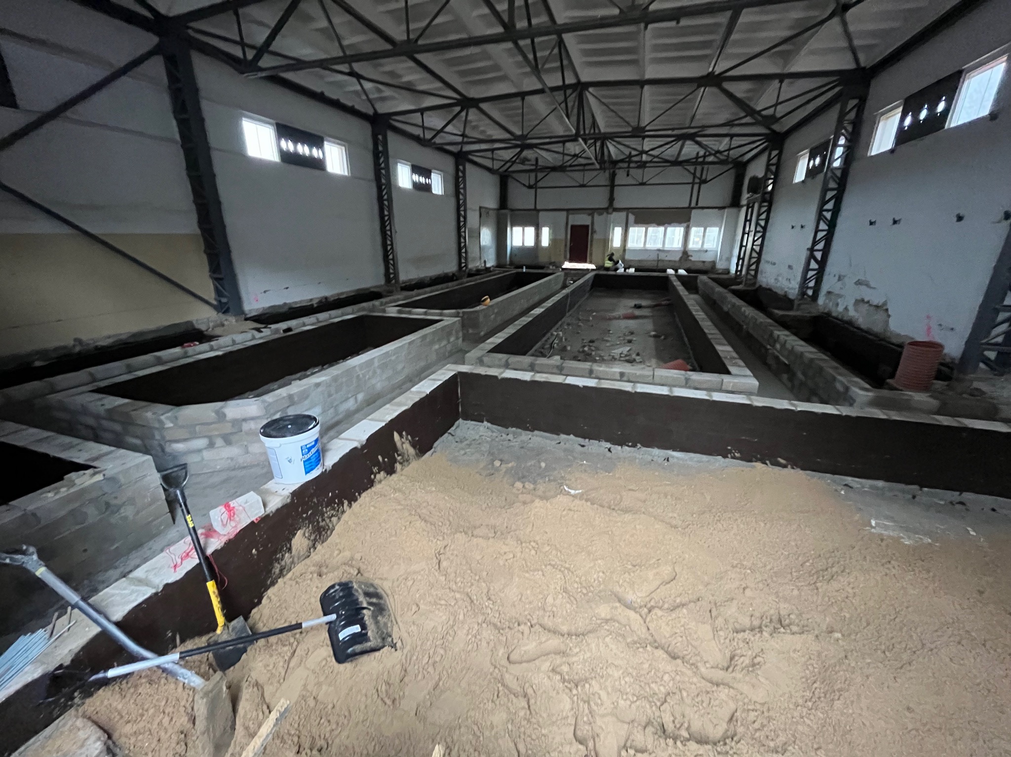 Wnętrze hali z przygotowanymi fundamentami pod budowę, widoczne narzędzia i materiały budowlane. Surowy stan, ceglane obrzeża, piasek.
