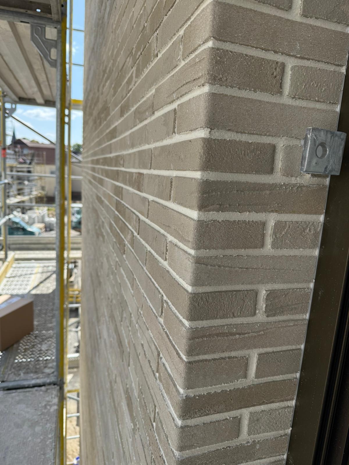 Ściana z beżowej cegły klinkierowej z białą fugą, widoczna z bliska, z fragmentem rusztowania po lewej stronie i metalowym elementem przy ścianie po prawej.