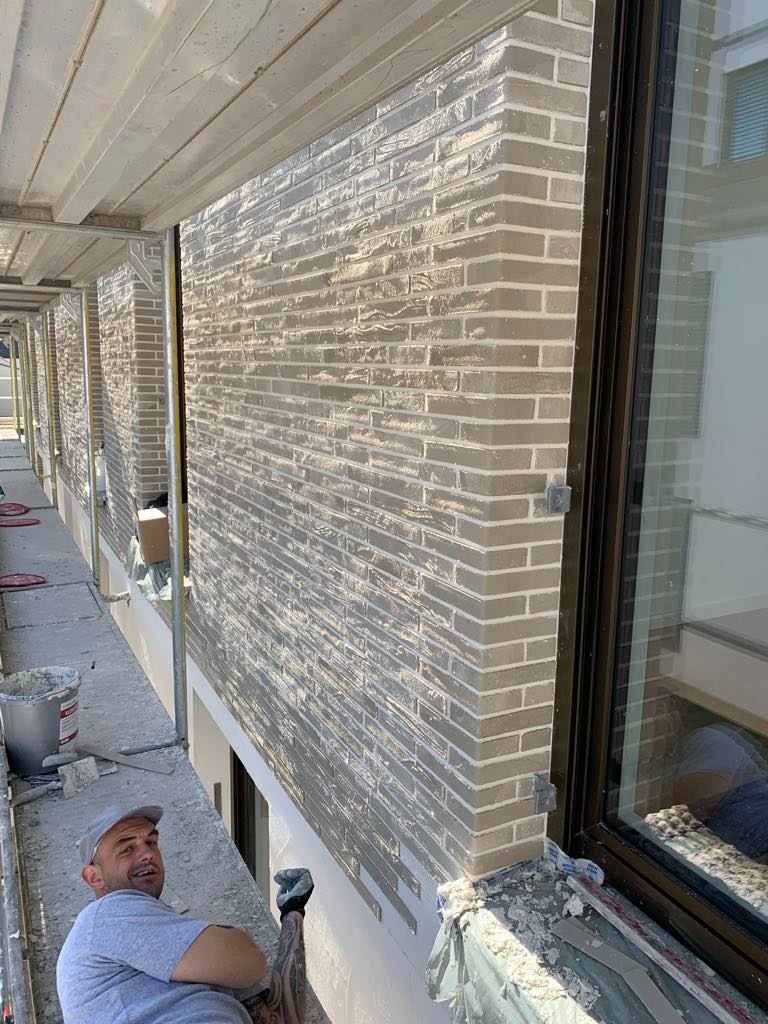 Ściana budynku wykończona cegłą klinkierową w odcieniach szarości, widoczna z bliska z rusztowania. Okno z brązową ramą. Praca w toku.