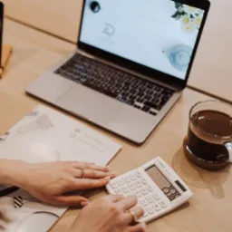 Kalkulator w dłoni, laptop z logo firmy, kawa i dokumenty na biurku. Scena z biura rachunkowego, sugerująca pracę z finansami i kontakt z klientem.