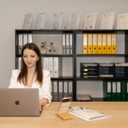 Kobieta w biurze pracuje na laptopie, za nią regał z segregatorami i certyfikatami. Na biurku kawa, telefon i dokumenty.