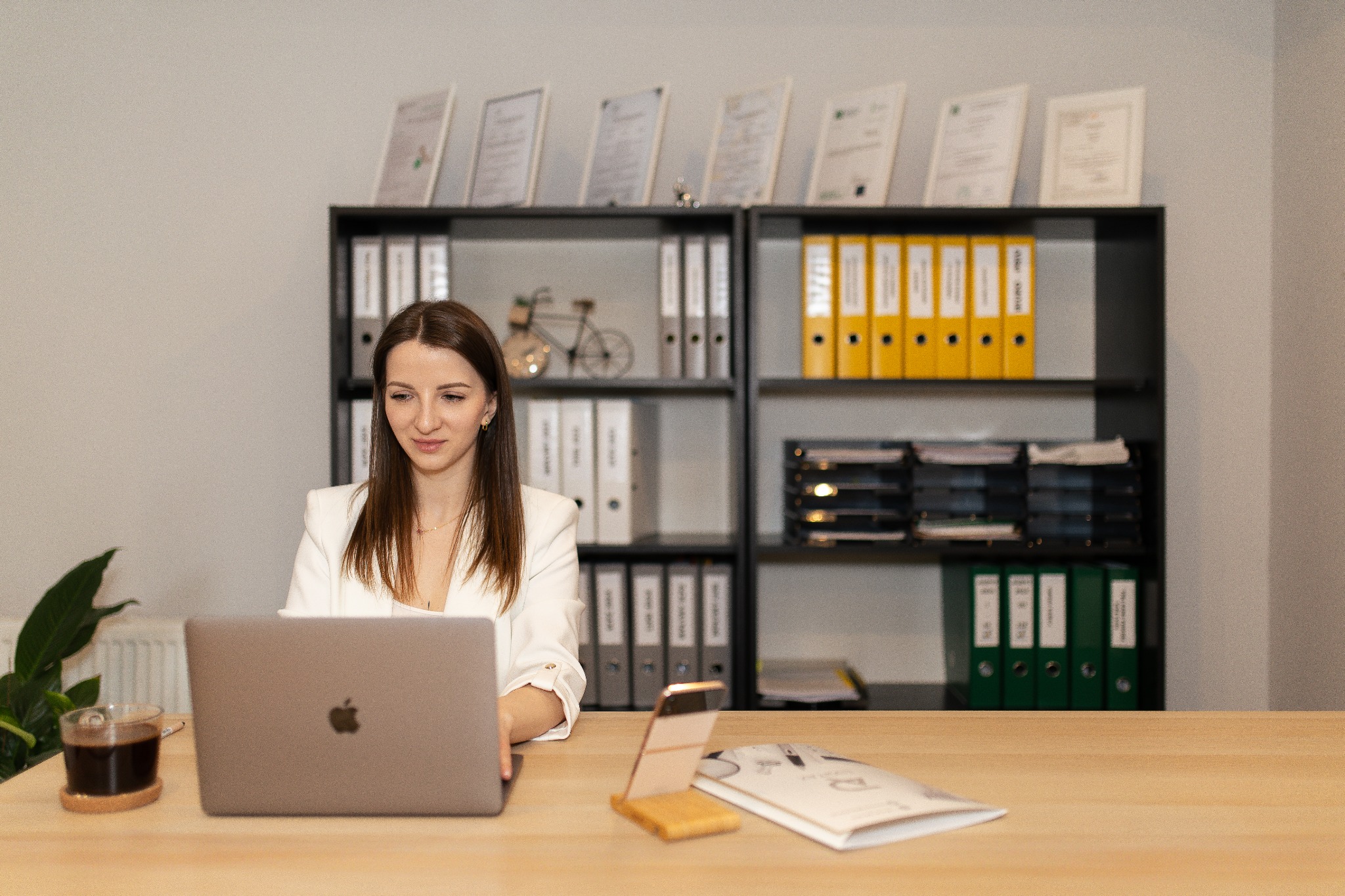 Kobieta w biurze pracuje na laptopie, za nią regał z segregatorami i certyfikatami. Na biurku kawa, telefon i dokumenty.