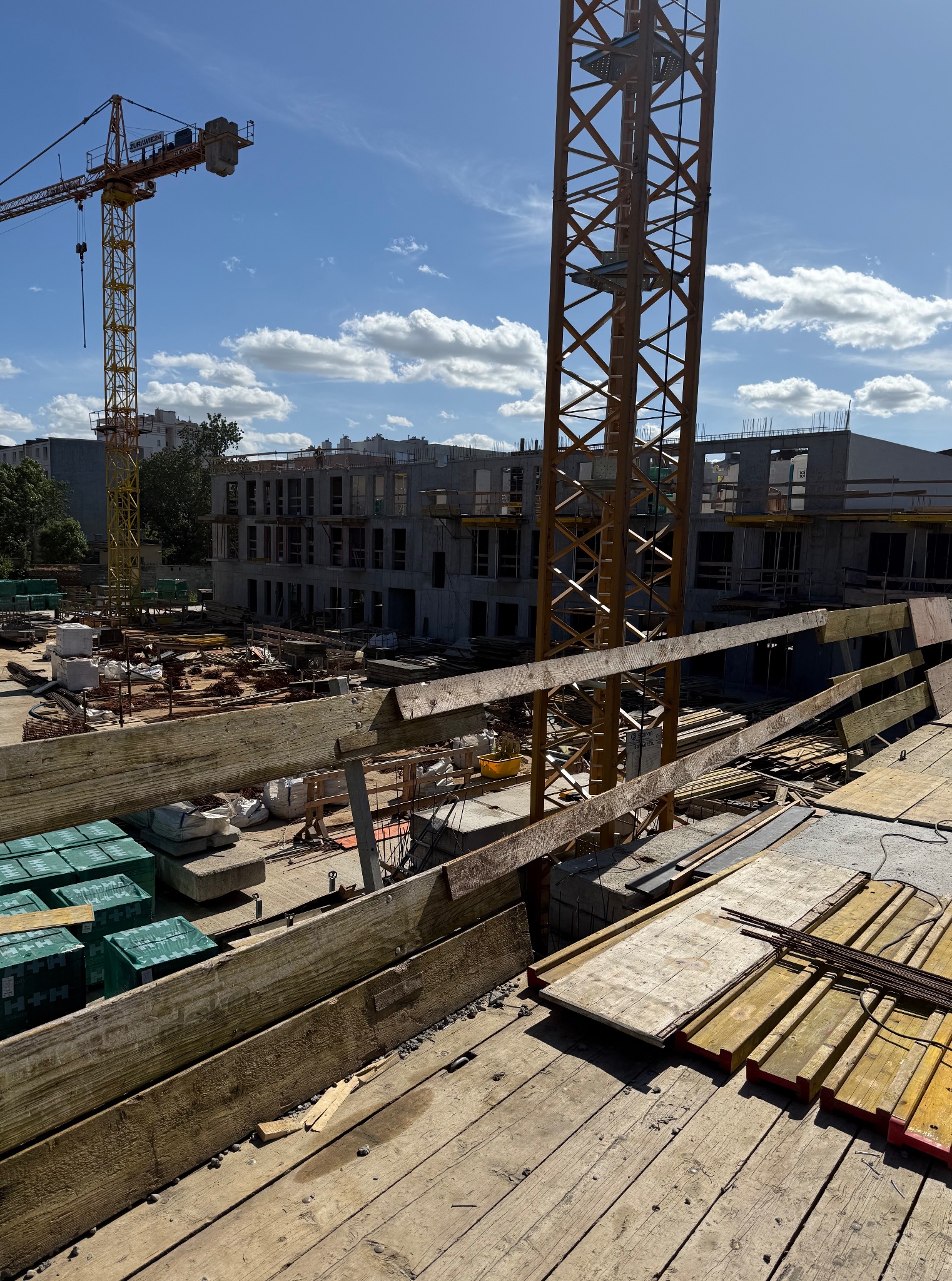 Budowa budynku w toku z widocznymi elementami konstrukcyjnymi, żurawiem i drewnianym szalunkiem. Słoneczny dzień, częściowo zachmurzone niebo w tle.