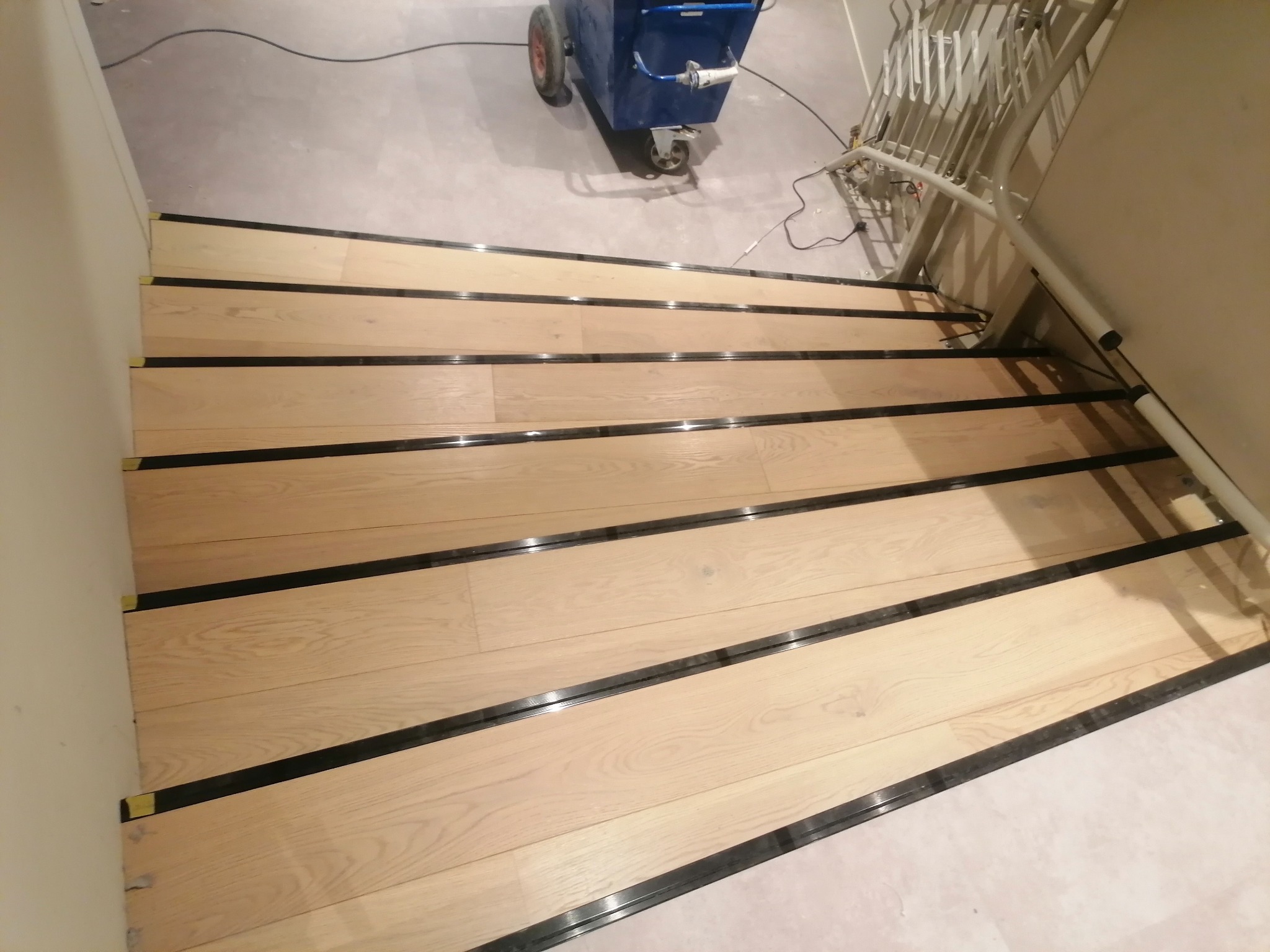 Jasne drewniane schody z czarnymi listwami, częściowo wykończone, z widoczną maszyną do czyszczenia podłóg w tle. Ujęcie z góry.