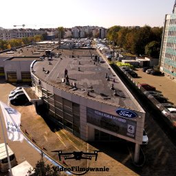 Videofilmowanie Anna Kusiak - Filmowanie dronem salonu Ford Auto Plaza z ekipą na dachu. Ujęcie z góry, widoczny dron, budynki w tle i zaparkowane samochody. Tekst 'VideoFilmowanie' na dole.
