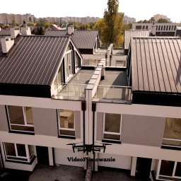 Videofilmowanie Anna Kusiak - Dron filmuje z góry nowoczesne, szeregowe domy z ciemnymi dachami i balkonami. Widoczne budynki w tle i fragment drzewa. Ujęcie z powietrza.