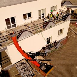 Videofilmowanie Anna Kusiak - Demontaż dachu budynku z użyciem rury zsypowej i kontenerów na gruz. Dwóch robotników w kaskach i kamizelkach na dachu. Widok z góry, z drona.