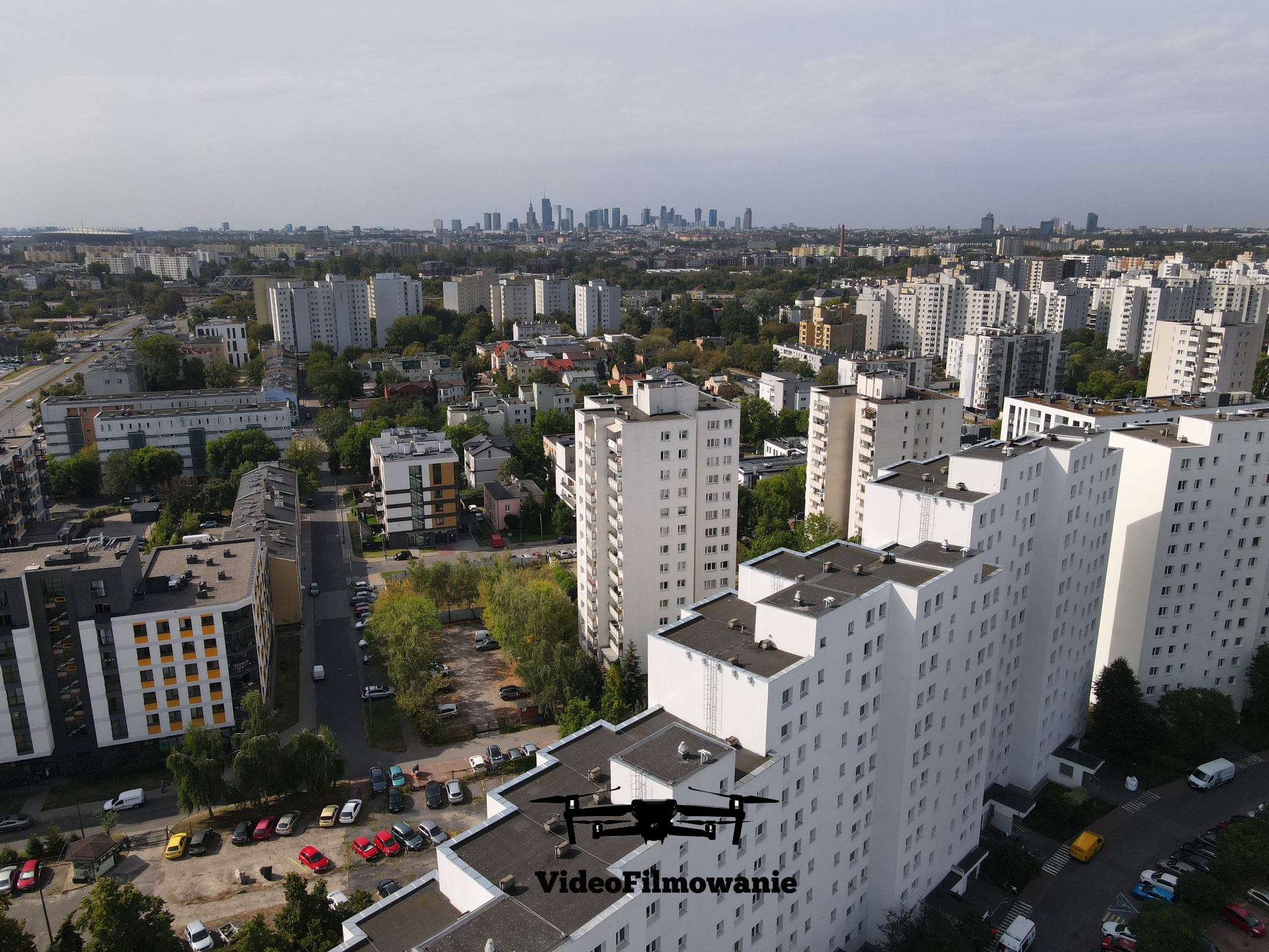 Zdjęcie z drona nad osiedlem mieszkaniowym z widokiem na panoramę miasta w tle. Na pierwszym planie sylwetka drona i napis 'VideoFilmowanie'.
