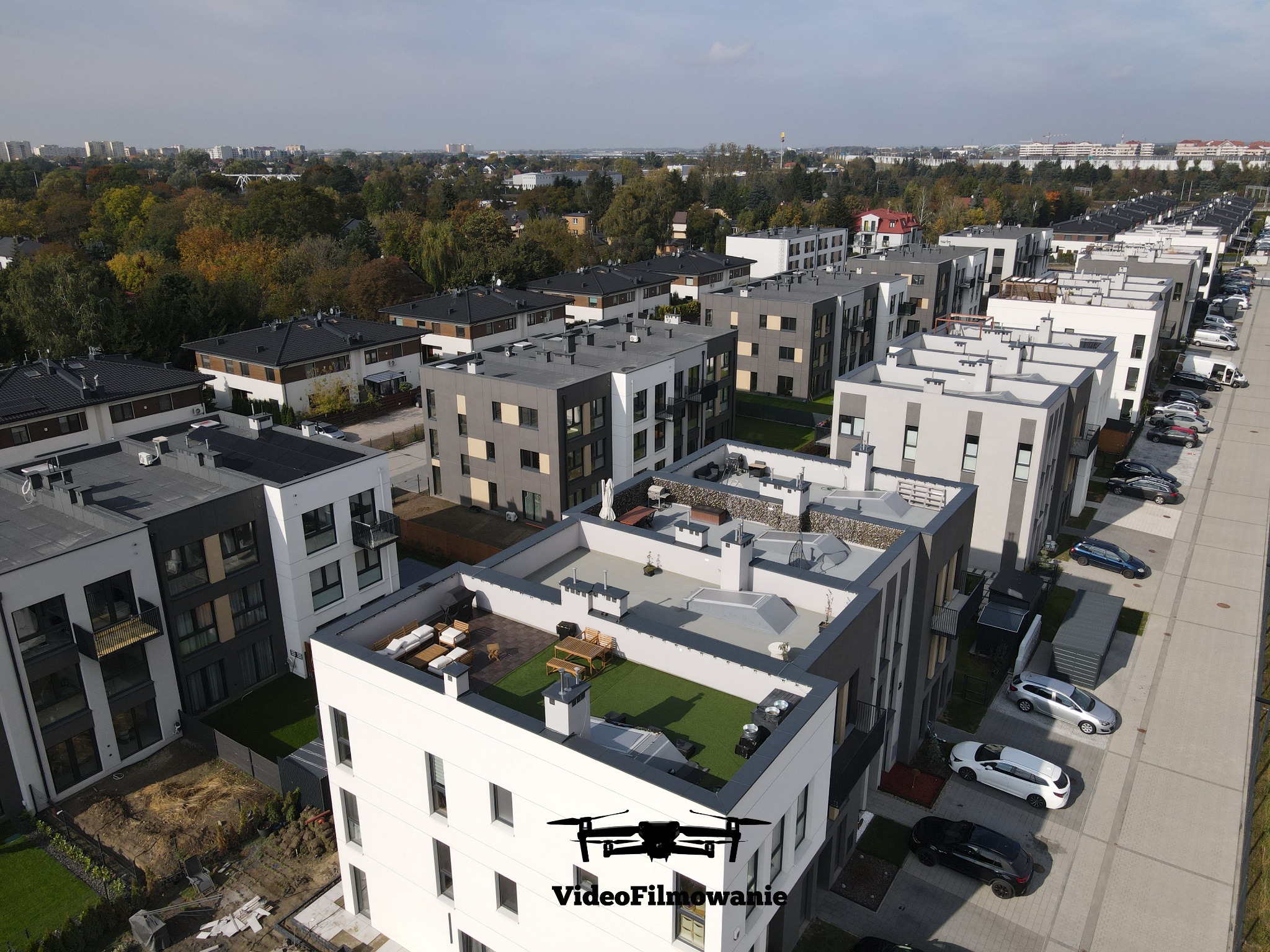 Zdjęcie z drona przedstawia osiedle nowoczesnych, wielopiętrowych budynków z płaskimi dachami i zielonymi tarasami. Widoczne zaparkowane samochody i drzewa w tle.
