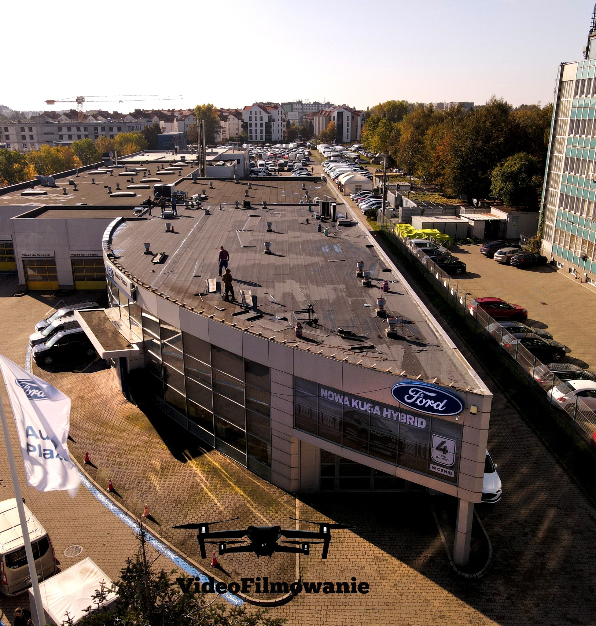 Filmowanie dronem salonu Ford Auto Plaza z ekipą na dachu. Ujęcie z góry, widoczny dron, budynki w tle i zaparkowane samochody. Tekst 'VideoFilmowanie' na dole.