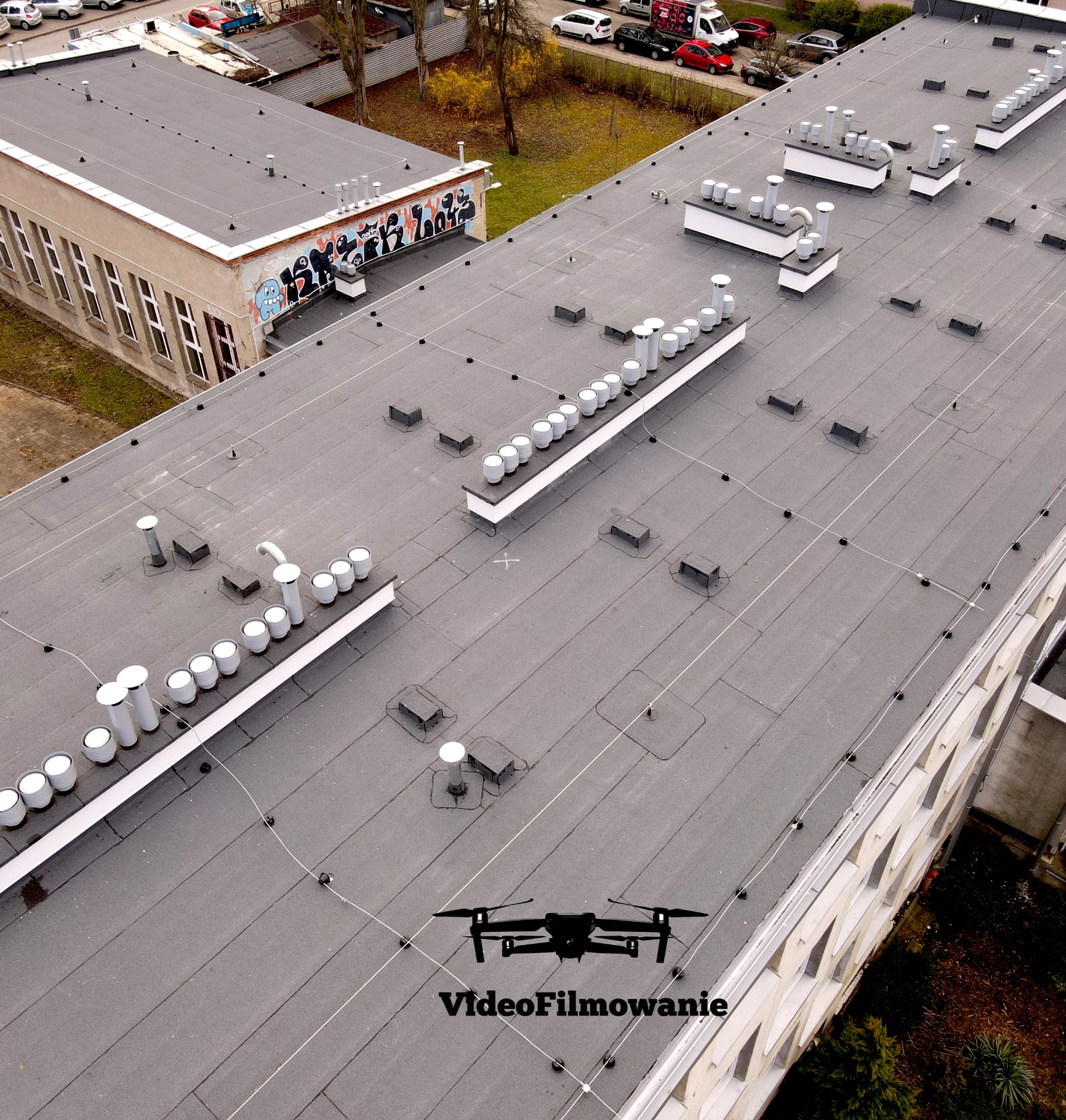 Zdjęcie z drona przedstawia płaski dach budynku z licznymi kominami wentylacyjnymi i instalacjami. Na dachu widoczne logo firmy 'VideoFilmowanie'.