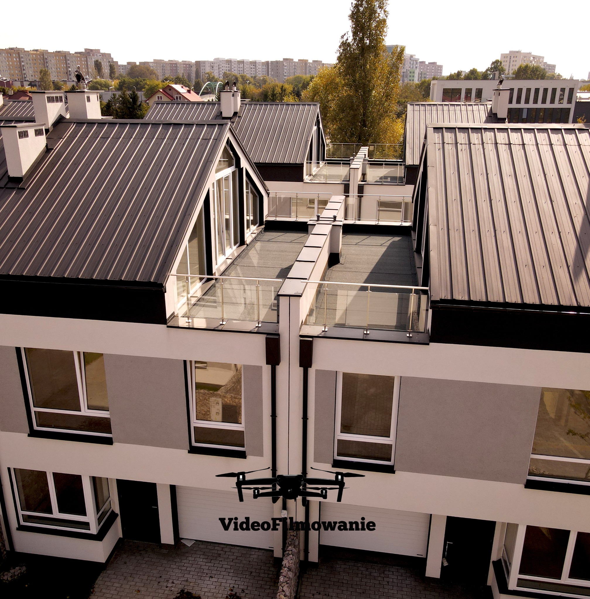 Dron filmuje z góry nowoczesne, szeregowe domy z ciemnymi dachami i balkonami. Widoczne budynki w tle i fragment drzewa. Ujęcie z powietrza.