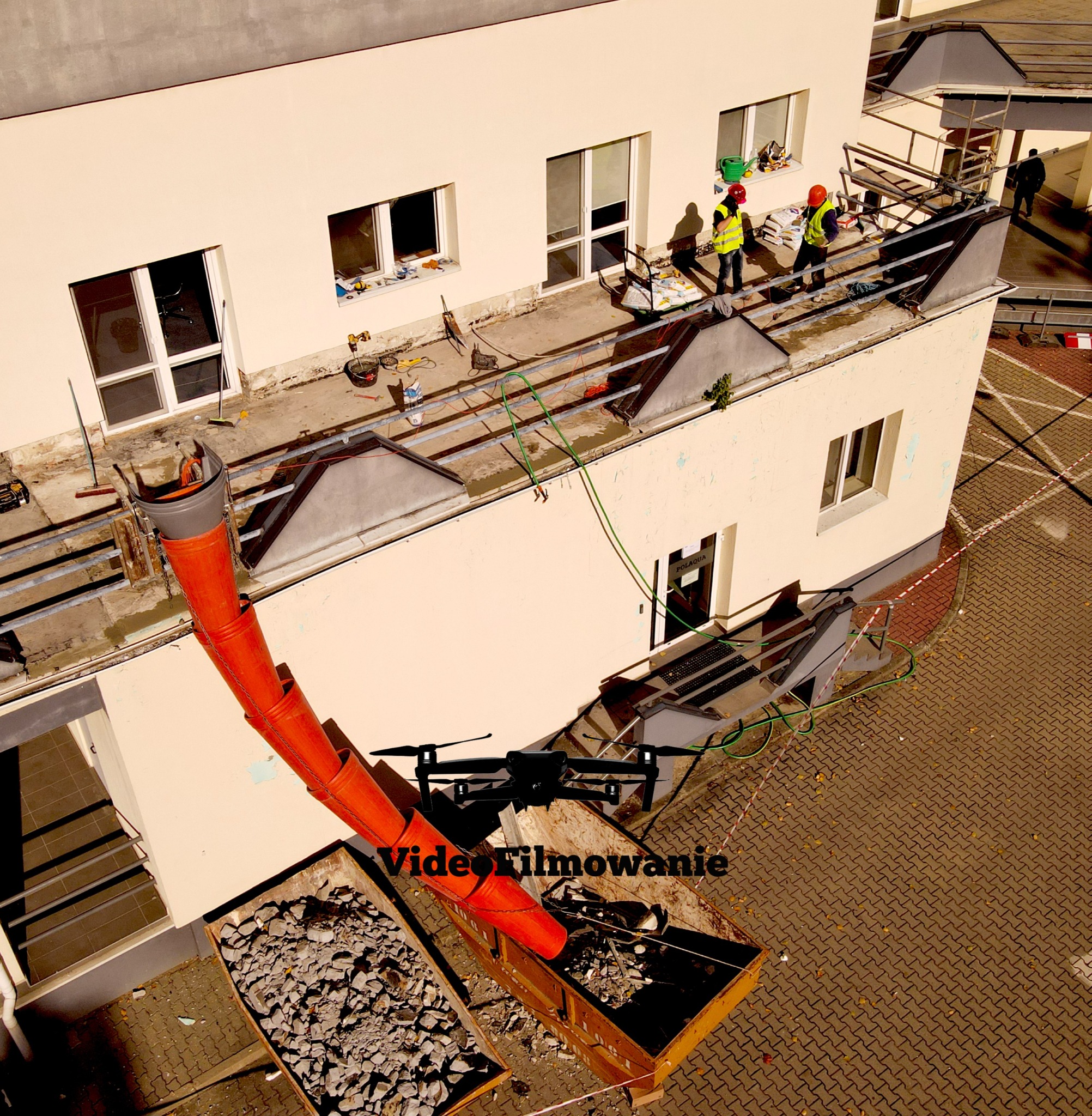 Demontaż dachu budynku z użyciem rury zsypowej i kontenerów na gruz. Dwóch robotników w kaskach i kamizelkach na dachu. Widok z góry, z drona.