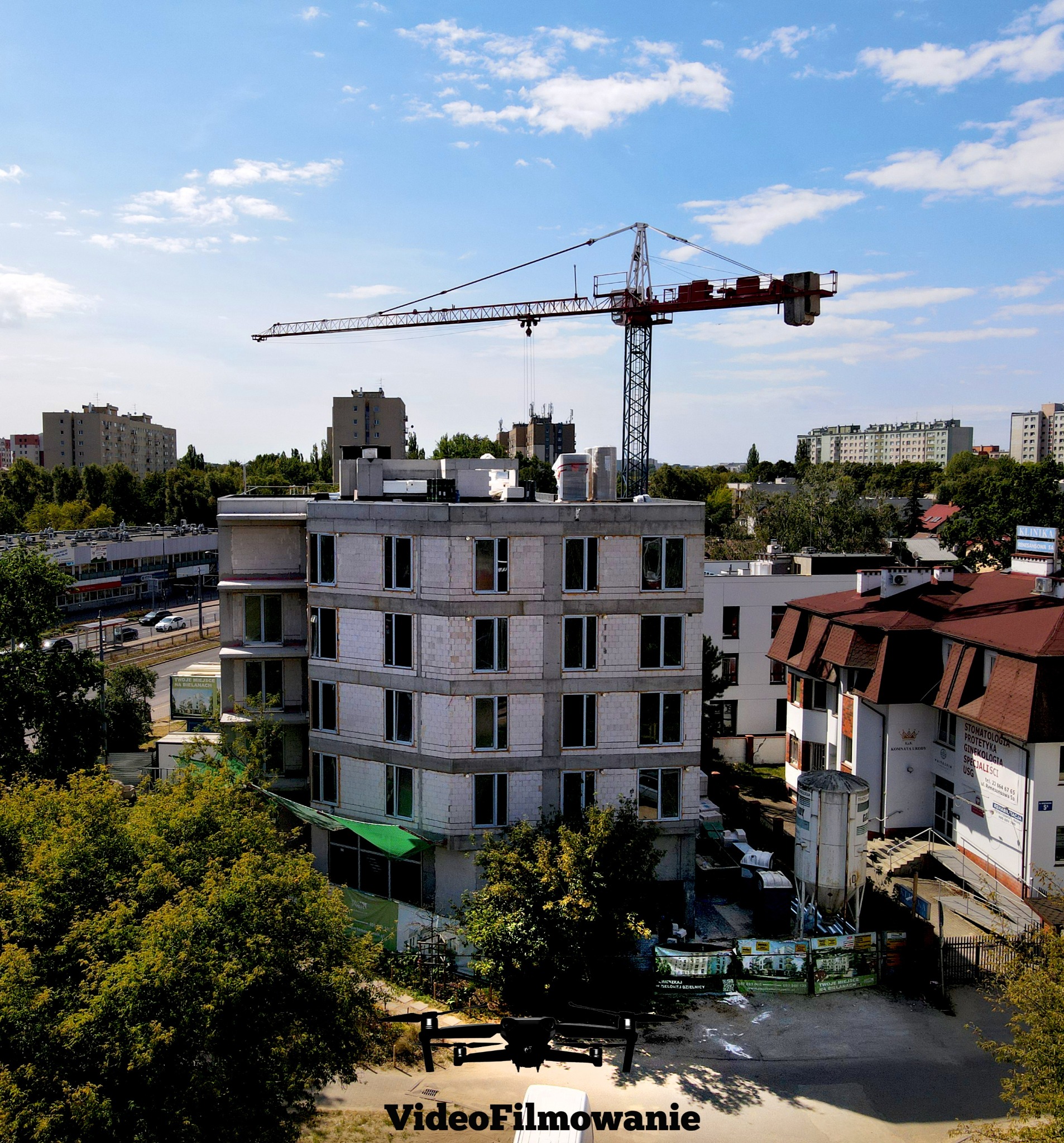 Budowa bloku mieszkalnego z żurawiem w tle, ujęcie z drona. Widoczne okoliczne budynki i zieleń miejska. Na dole kadru cień drona.