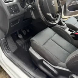 Wnętrze białego Citroena C4: czarna tapicerka fotela kierowcy, dywaniki, deska rozdzielcza i kierownica z logo. Widoczne pedały i dźwignia regulacji fotela.